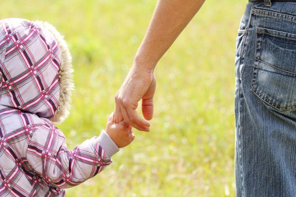 A man holds the hand of his young daughter as they walk through a field.