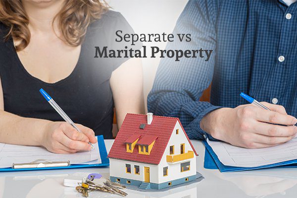 A man and a woman sitting beside each other signing papers with a mini house figure and a set of keys between them and the words separate vs marital property